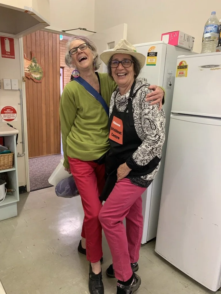 Two elderly women with gray hair and glasses are laughing and hugging in a kitchen. One woman wears a green sweater, pink pants, and black shoes, while the other wears a hat, a patterned hoodie, pink pants, and a black apron with a welcome badge. The