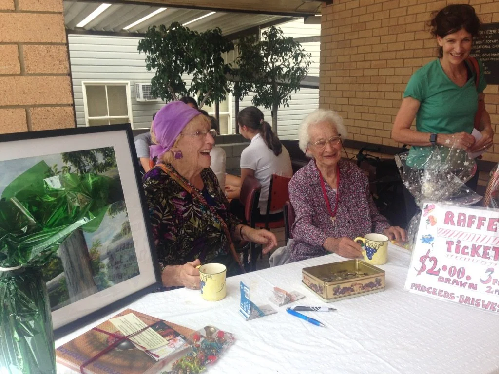Two elderly women sitting at a table with a framed photo, cups, and raffle ticket sign, smiling and talking. A woman standing behind them is holding a gift bag, in an indoor setting with green plants and other people in the background.