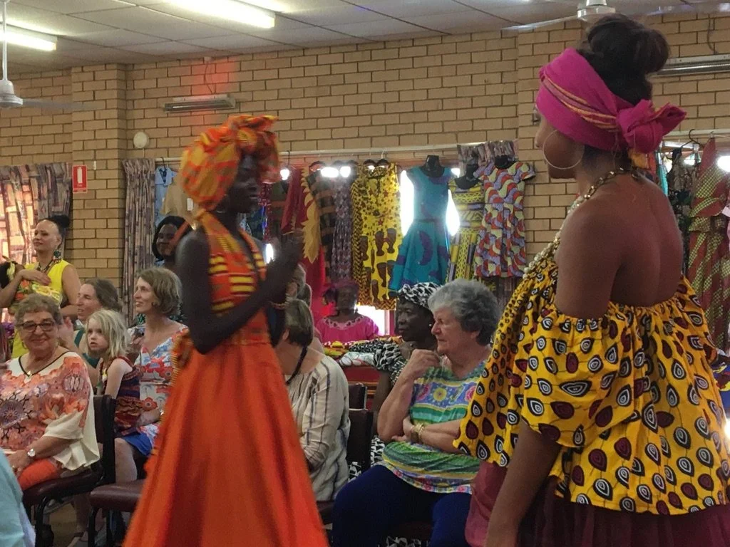 Two women in traditional African clothing are standing in front of an audience. The woman on the right is wearing a yellow dress with blue and red patterns, a pink head wrap, and large hoop earrings. The woman on the left is dressed in an orange dres