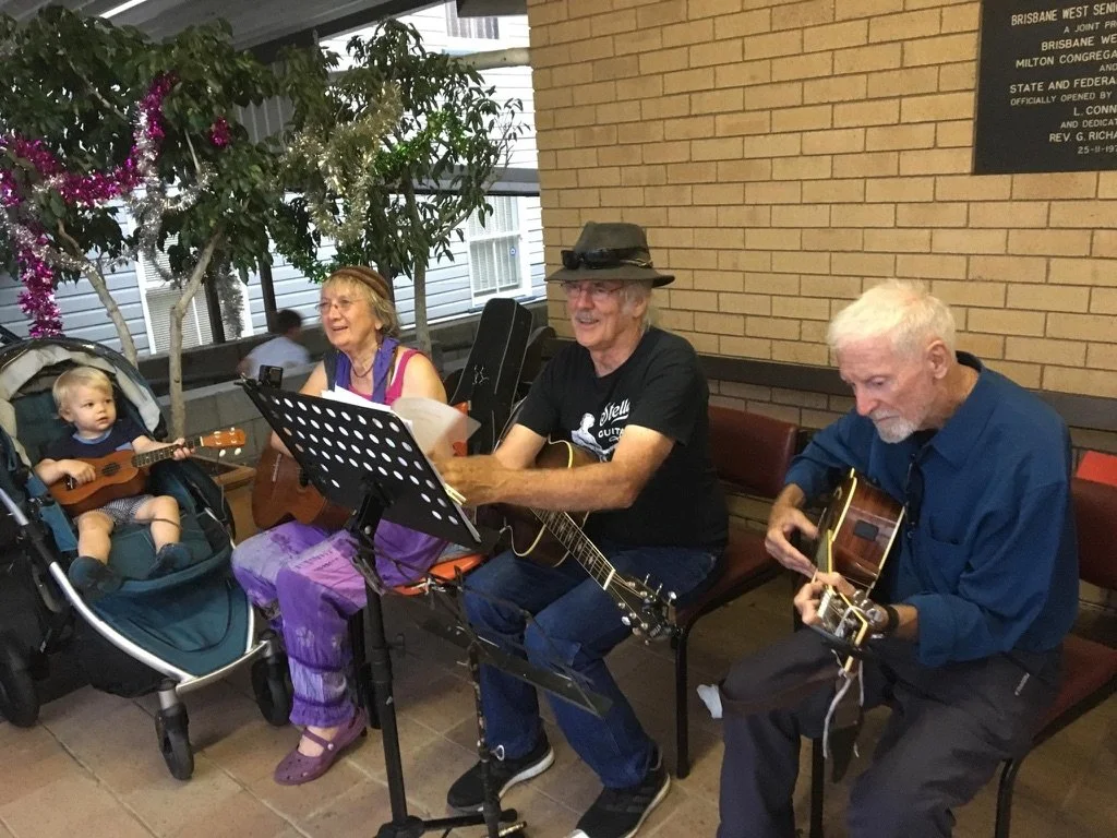 Two elderly men and a woman are playing guitars and singing, while a young girl sits in a stroller nearby, holding a small guitar. They are seated indoors with a brick wall and decorative trees in the background.