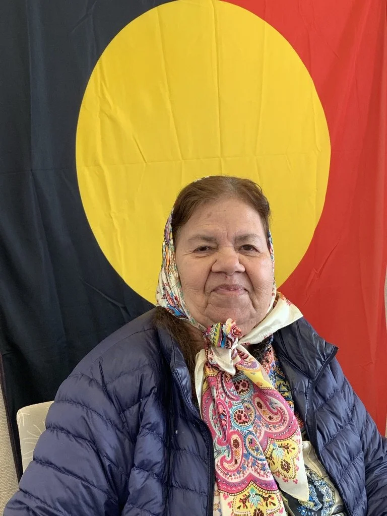 A woman wearing a colorful scarf and a navy blue jacket, sitting in front of a flag with a yellow circle on a black and red background.