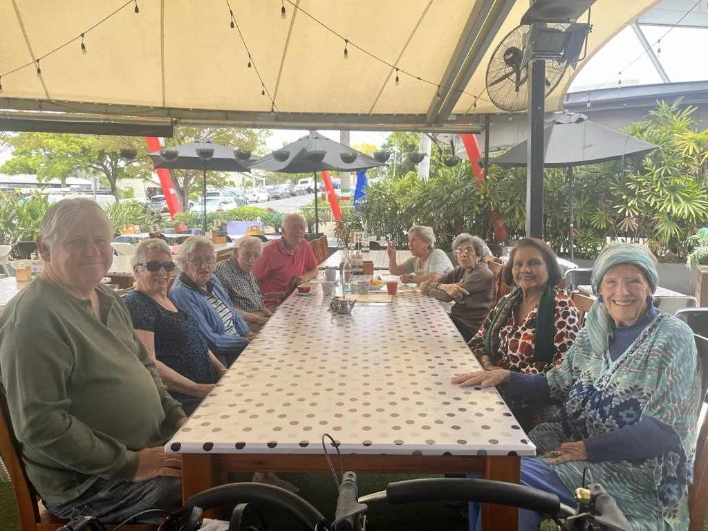 A group of elderly people sitting around a long table at an outdoor restaurant or patio, under a canopy with string lights and surrounded by greenery.