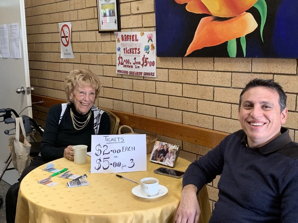 An elderly woman and a middle-aged man sitting at a table selling raffle tickets, with a sign indicating $2 each or 3 for $5, and a no smoking sign on the brick wall behind them. The woman has a walker beside her, and there are coffee cups and miscel