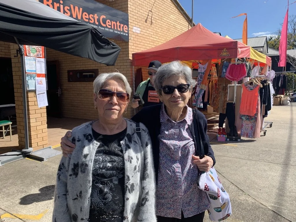 Two elderly women standing outside a community center with a market stall behind them selling clothing and accessories. One woman is wearing sunglasses, a black and white top, and a light gray cardigan, while the other is wearing sunglasses, a patter