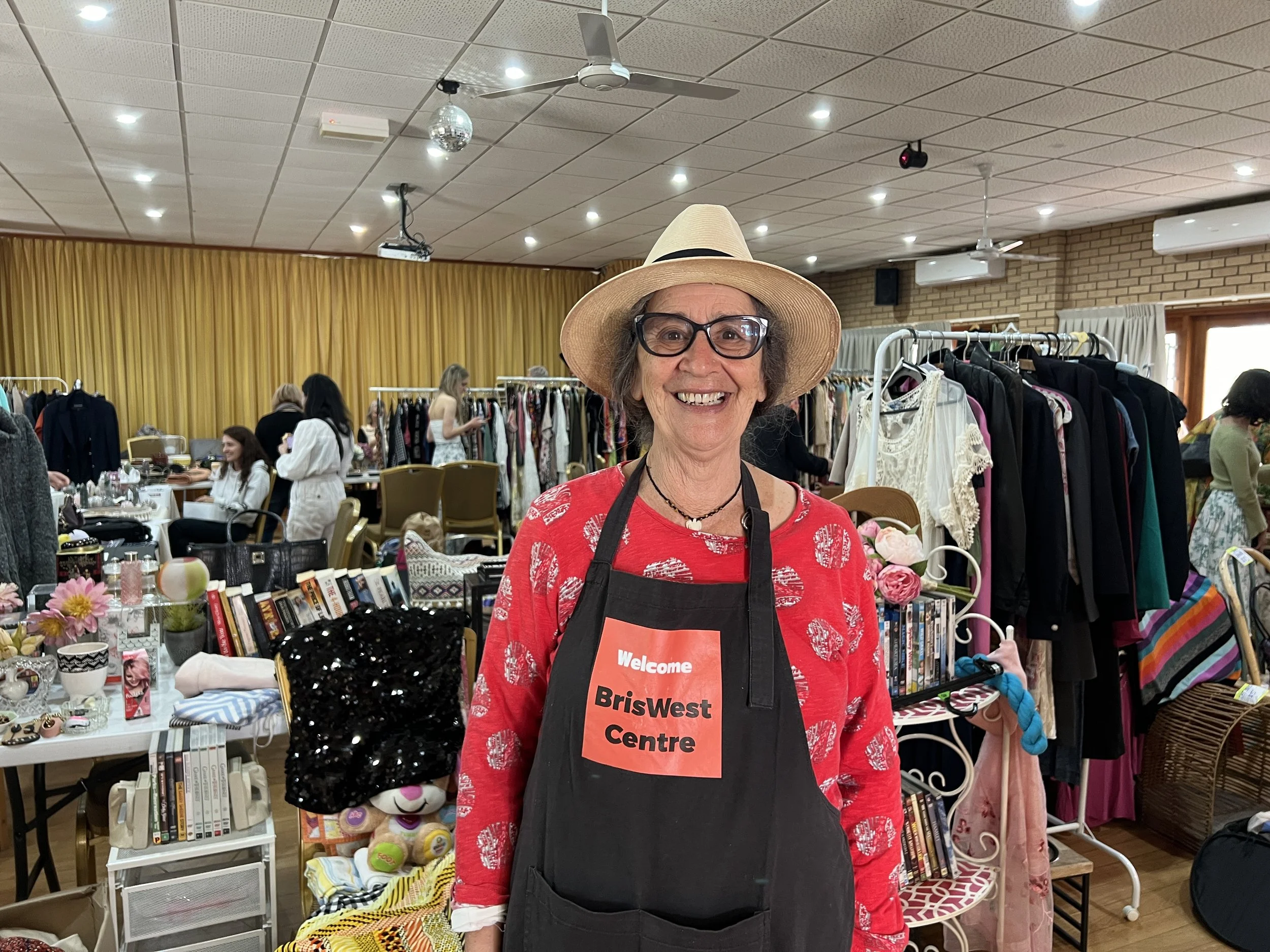 A smiling woman wearing a straw hat, black glasses, and a black apron with a red welcome sign, standing in a room filled with clothes and accessories for sale at a thrift store or sale event.