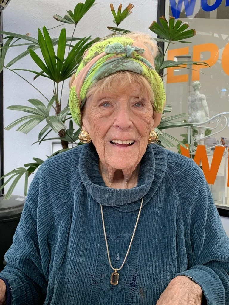 An elderly woman wearing a colorful headscarf, large gold earrings, a blue sweater, and a necklace, smiling at the camera with plants and a window with orange and blue writing in the background.