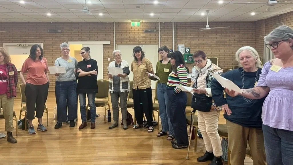 Group of women standing in a line in a community room, holding papers and participating in a group activity or discussion.