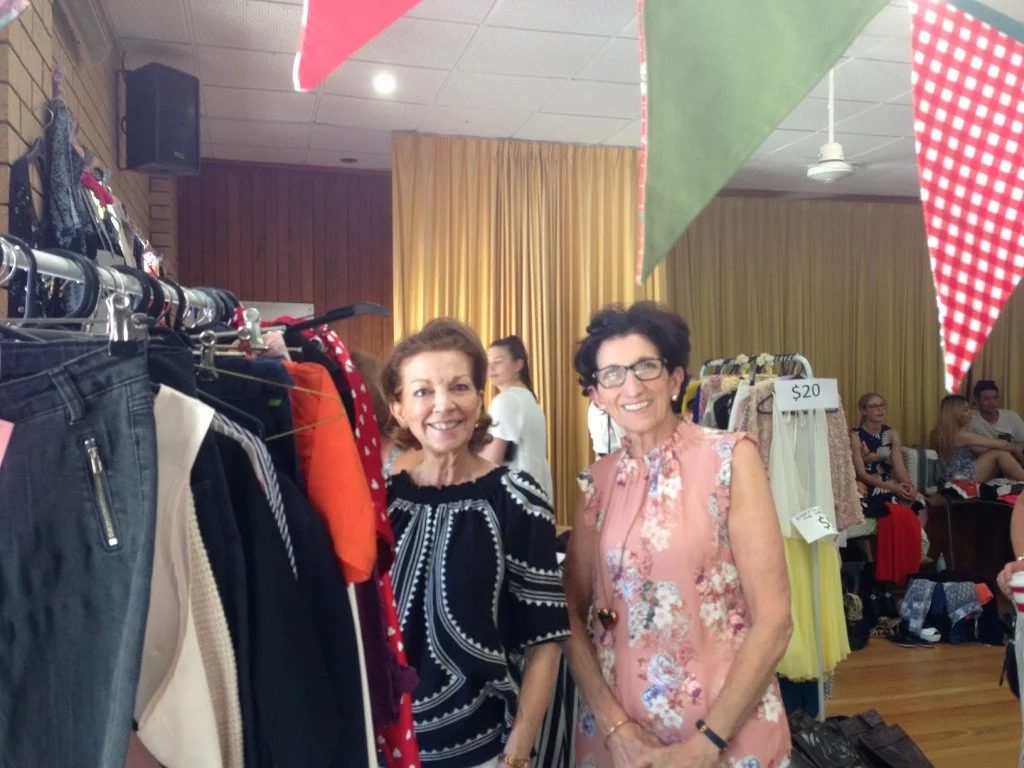 Two women shopping at a thrift store or flea market, standing next to a rack of clothes with a sign that says $20, with other shoppers seated in the background.