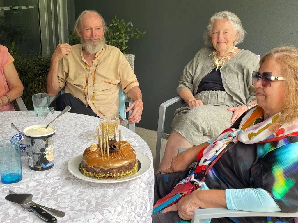 Group of four elderly people sitting around a table with birthday cake and drinks, celebrating on a patio or balcony.