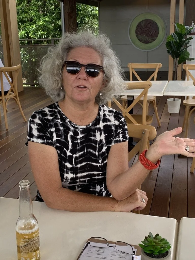 An older woman with gray curly hair wearing sunglasses, a black and white patterned shirt, and a red bracelet, sitting at an outdoor patio table. She is gesturing with her right hand while speaking. There is a bottle of beverage, a pair of glasses, a