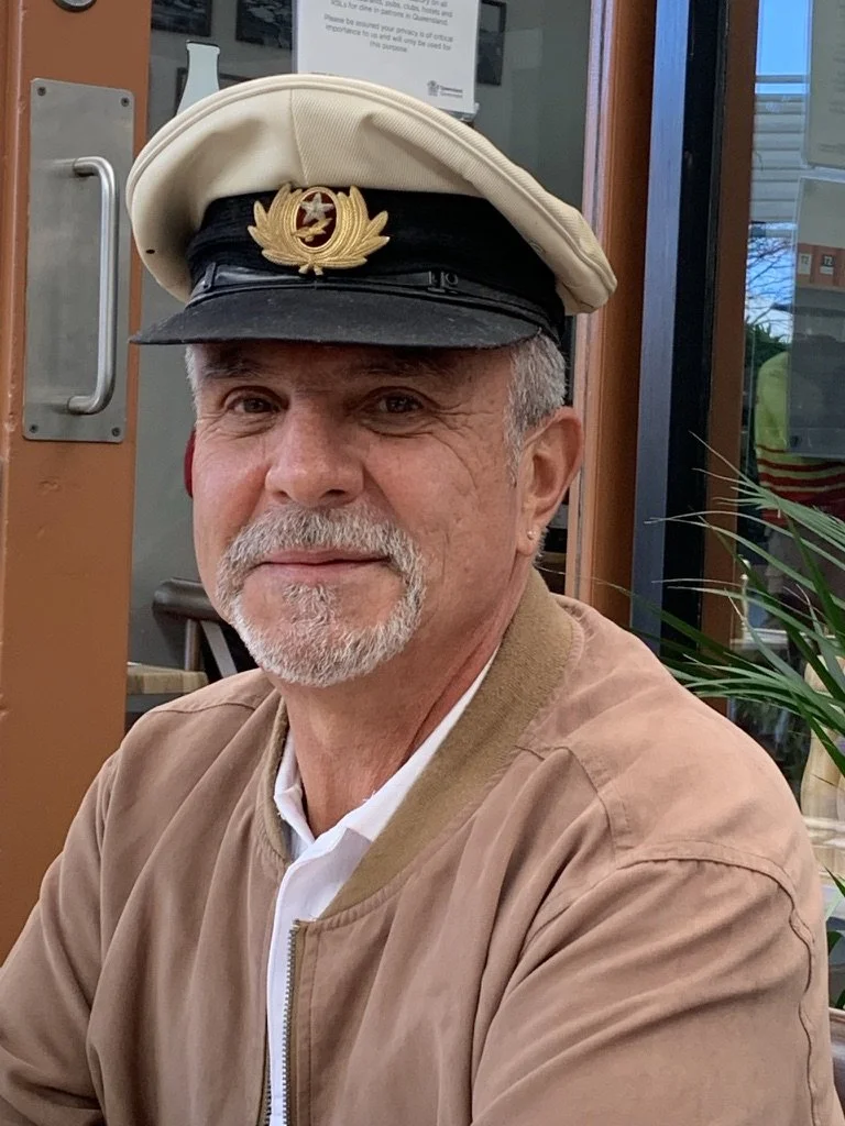 A man wearing a vintage military-style cap with a badge, a beige jacket, and a white shirt, sitting at a table indoors.