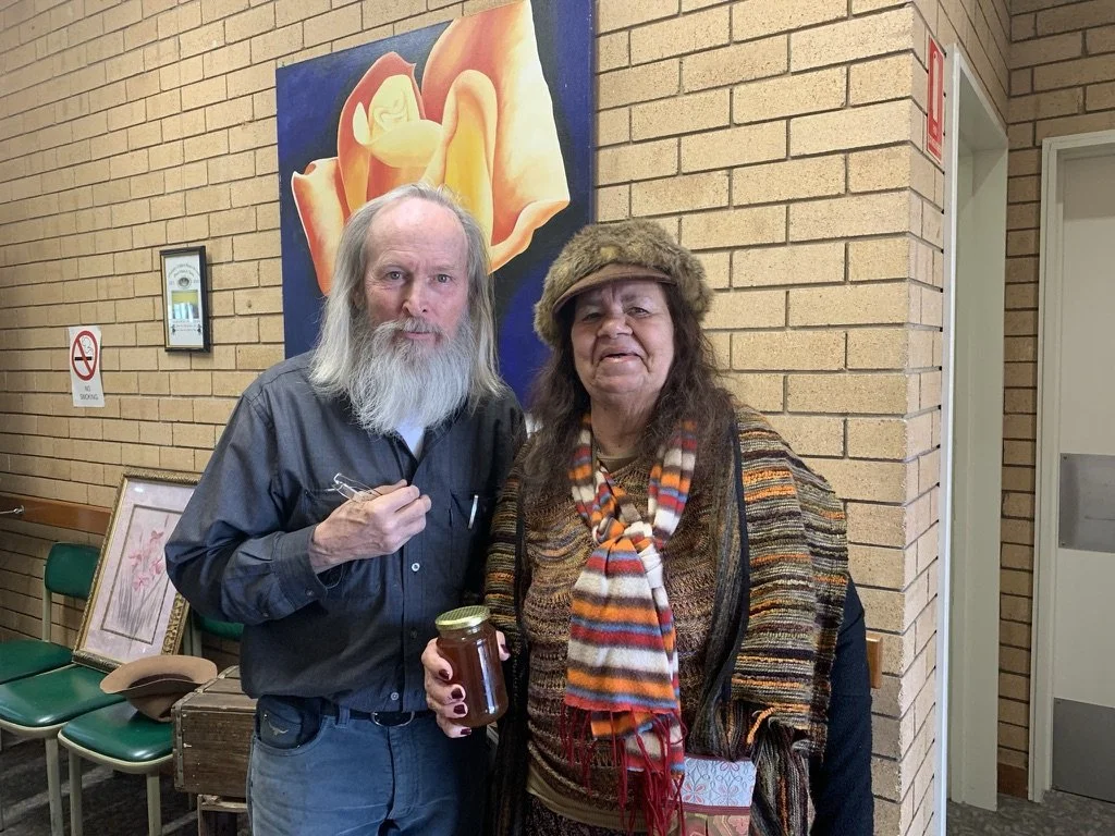 A bearded man wearing a dark shirt and a woman with long hair wearing a fur hat and colorful clothing holding a jar in a room with brick walls and a large colorful painting of a flower in the background.