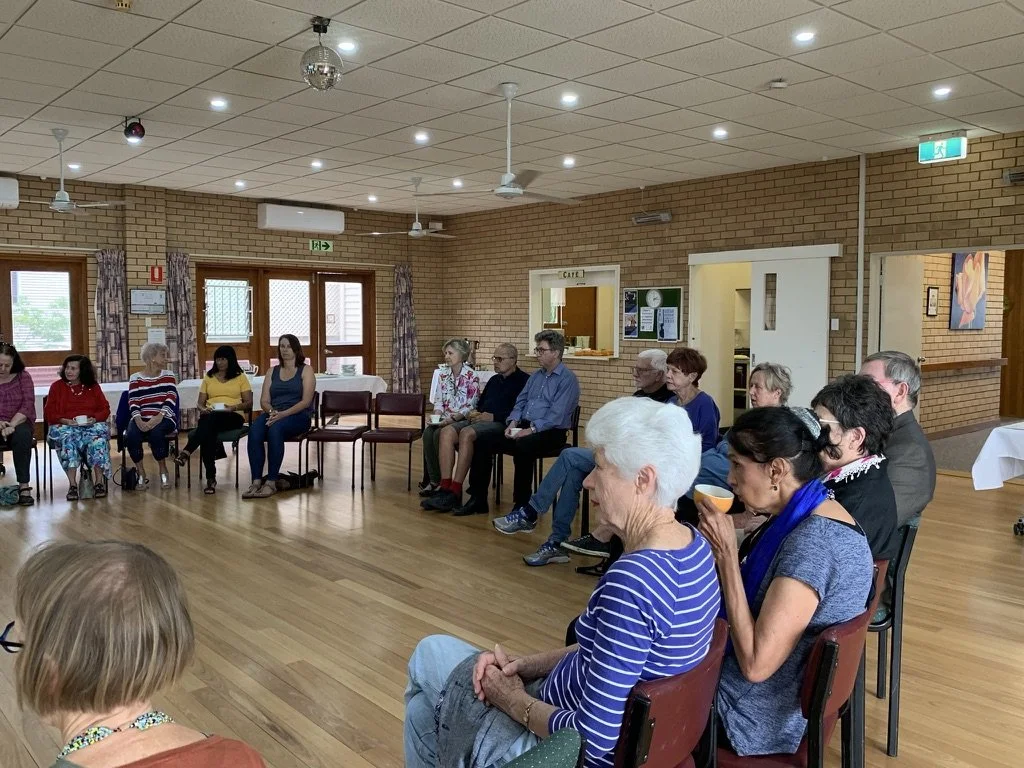 A group of people sitting in a circle inside a room, likely attending a meeting or gathering.