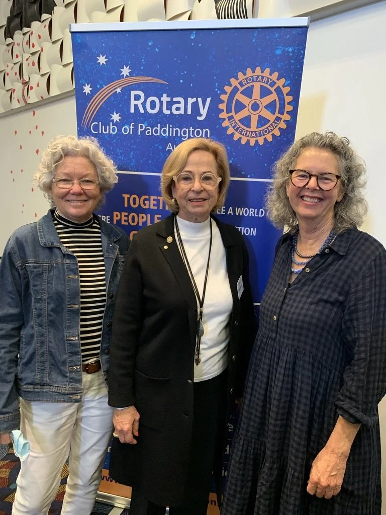 Three women standing in front of a Rotary Club of Paddington banner at a club event.