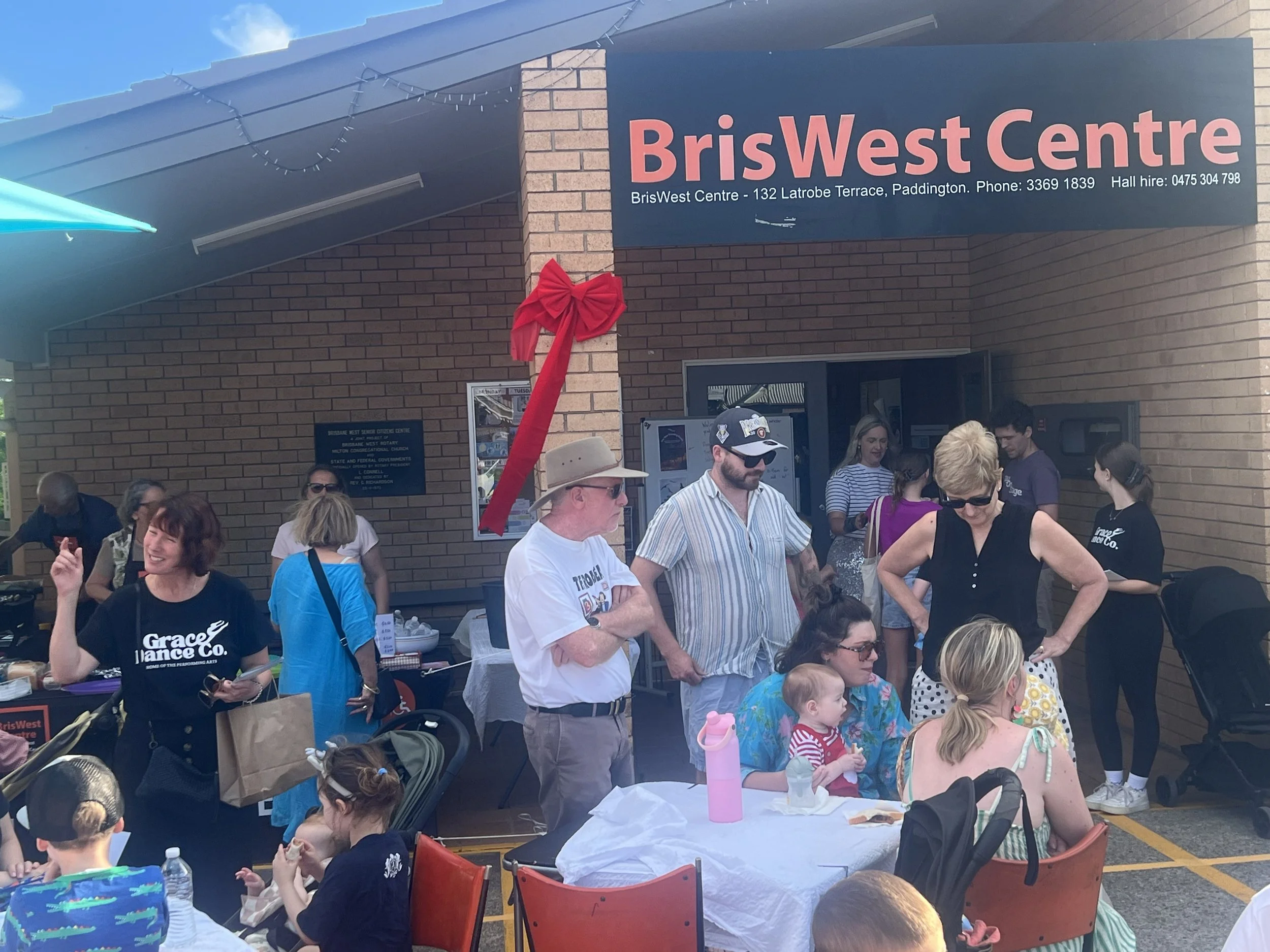 People gathered outside the BrisWest Centre during a daytime event, some seated at tables and others standing, with a large red bow decoration on the building.