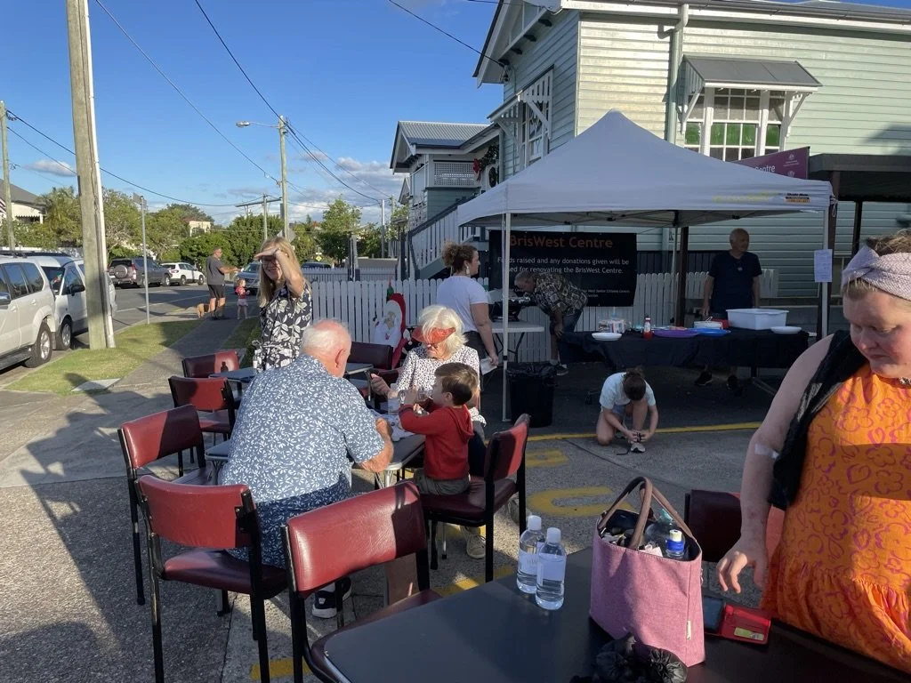 People gathered outdoors at a community event near a light green wooden building with a white picket fence. Some are seated at tables, while others are standing or crouching near a white canopy tent.