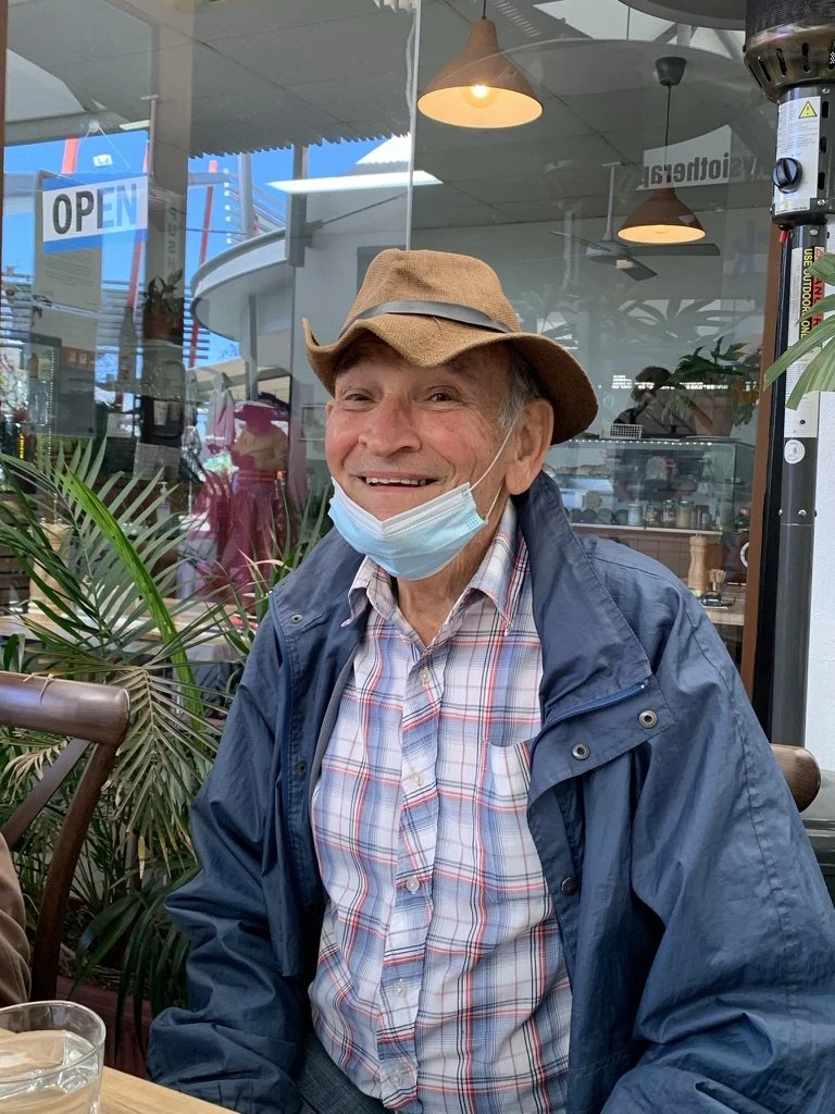 Smiling elderly man wearing a face mask, plaid shirt, and brown hat, sitting at a table in a restaurant or cafe with indoor plants and glass windows.