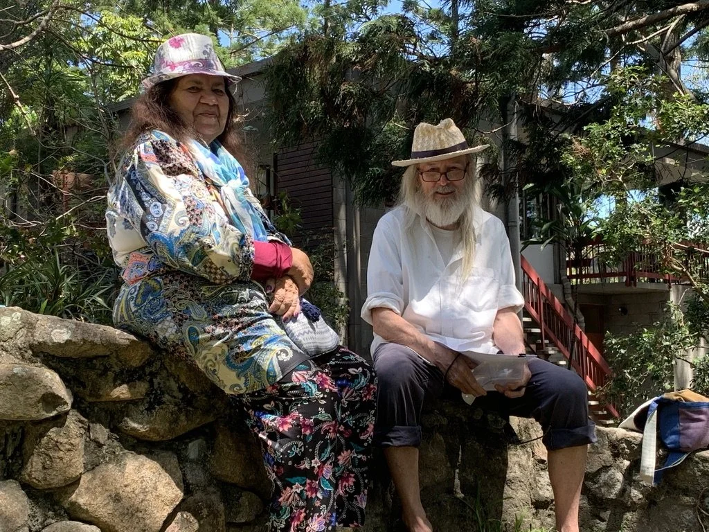 Two elderly people sitting on a stone wall outdoors, surrounded by trees and greenery. The woman on the left wears a colorful, patterned dress, pink gloves, and a pink hat. The man on the right has a long white beard, glasses, a straw hat, and a whit