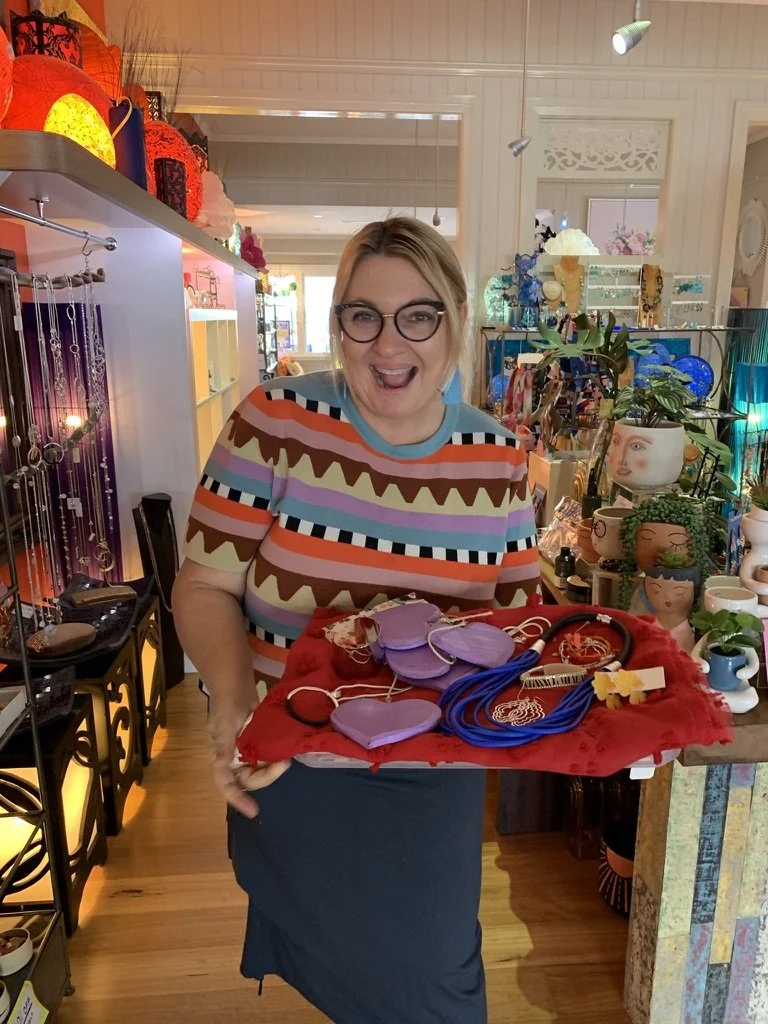 A woman with blonde hair and glasses smiling while holding a red cloth-covered tray with purple and pink jewelry in an arts and crafts store or boutique.