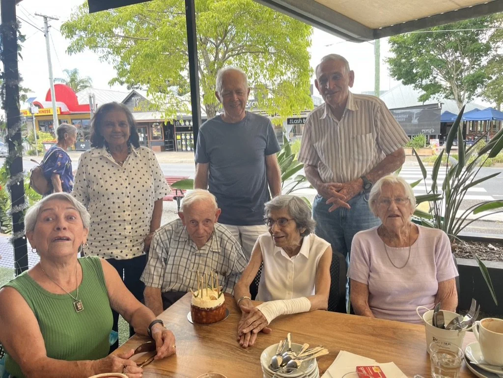 Group of seven elderly and middle-aged people celebrating a birthday at a restaurant, with one person holding a small birthday cake with candles, seated around a table with cups and utensils.