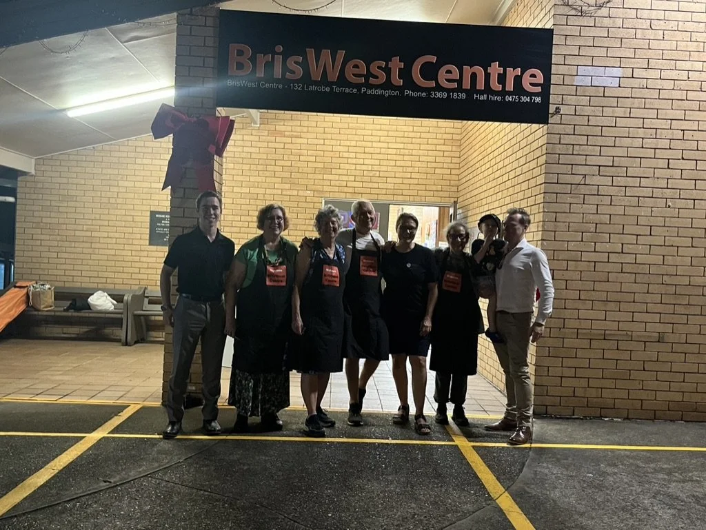 Group of seven people standing outside the BrisWest Centre at night, posing for a photo under a sign, with a brick wall backdrop and a small orange table to the side.