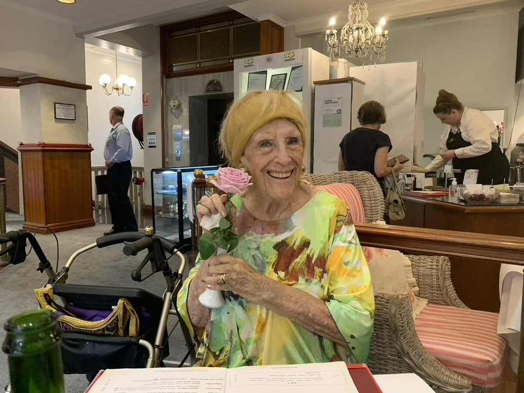 An elderly woman with a yellow headscarf and a colorful dress holding a pink rose inside a restaurant or café. She is smiling, sitting at a table with an open menu. In the background, there are staff members and a man standing near the wall.