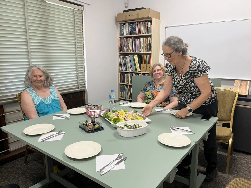Three women gathered around a table in a room with bookshelves. One woman is standing and serving food, while the other two sit and smile.