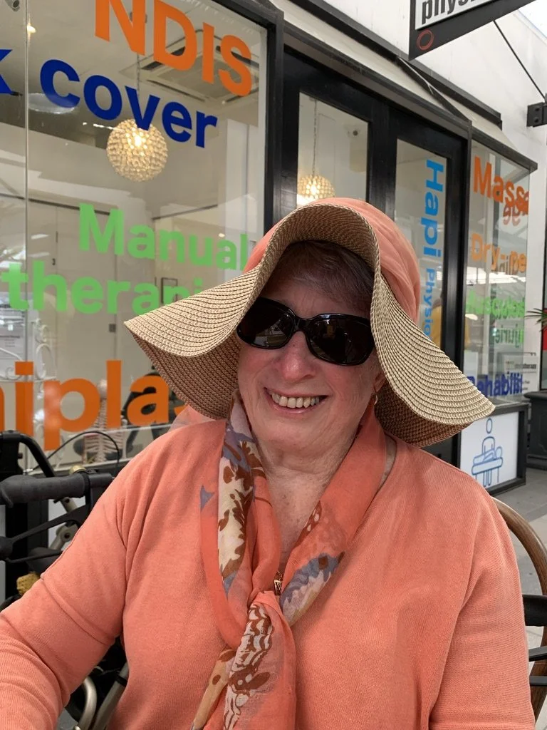Smiling older woman wearing a wide-brimmed sunhat, sunglasses, and an orange shirt with a colorful scarf, sitting outside a glass storefront with colorful signage.