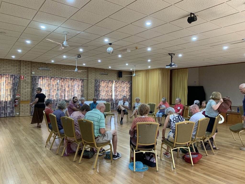 A group of elderly people sitting in a circle in a community hall with wooden floors, brick walls, and patterned curtains, participating in an indoor activity or meeting.
