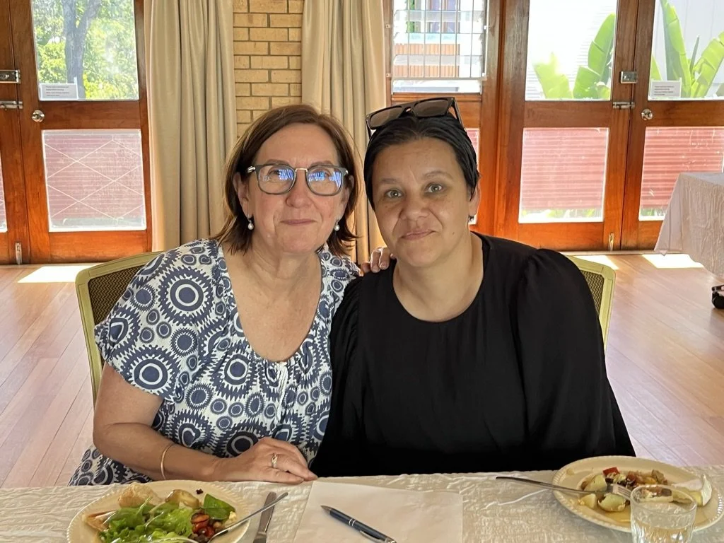 Two women sitting at a table with plates of food, indoors with wooden doors and windows in the background.