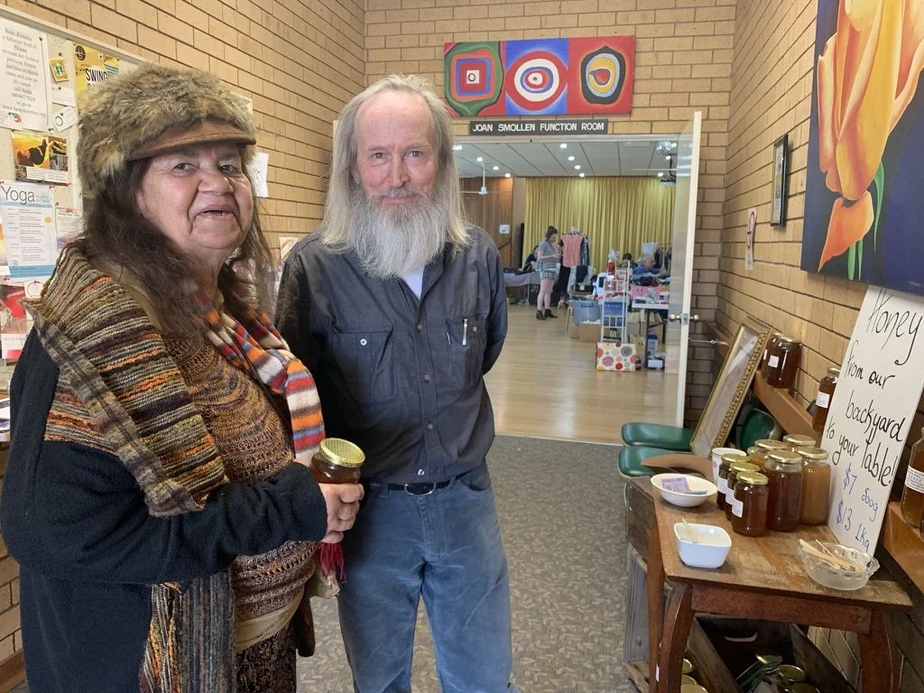 Two elderly individuals, a woman and a man, stand inside a community hall or event space. The woman is holding a jar of honey, wearing a colorful scarf, hat, and layered clothing. The man has long gray hair and a beard, wearing a dark shirt. Behind t