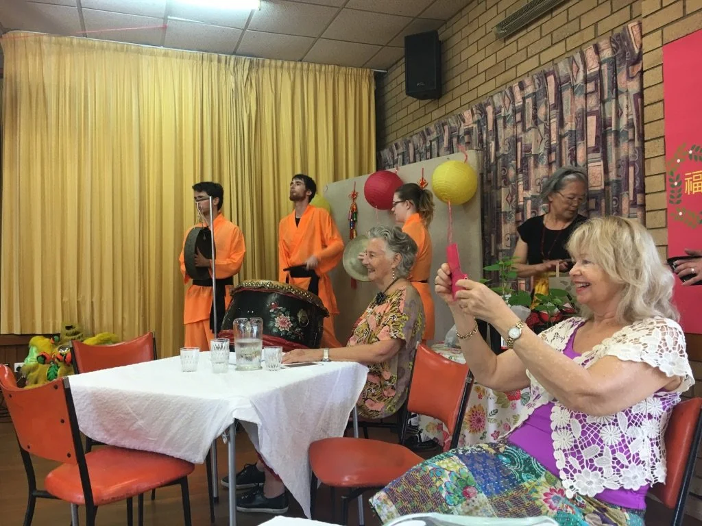 Group of women sitting at a table, one taking a photo with her phone, with performers dressed in orange costumes and traditional decorations in the background.