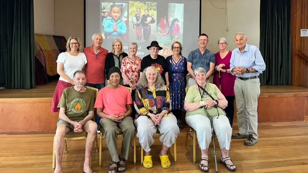 Group of eleven people, seven women and four men, gathered on a stage in an indoor setting. Some are seated on chairs in front, and others are standing behind. A projected screen displaying a collage of photos is visible in the background. The room h