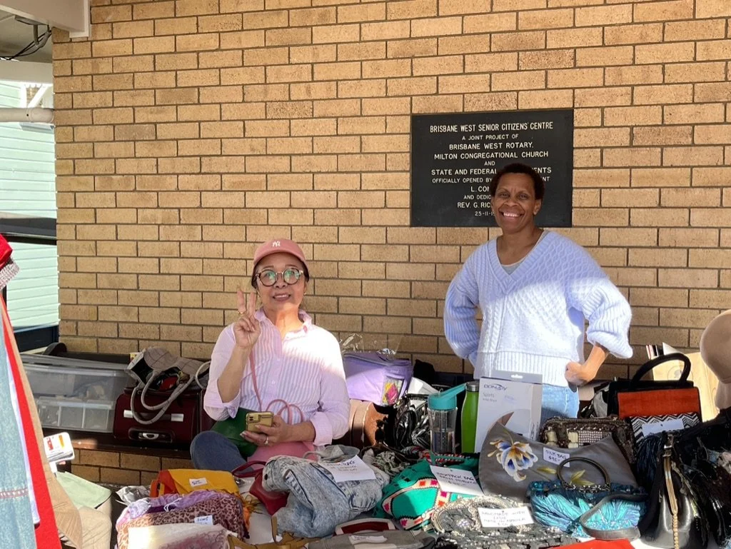 Two women standing behind a table filled with various items at an outdoor event. One woman is wearing glasses, a pink hat, and a striped shirt, holding a gold phone, while the other is wearing a white sweater and smiling. There is a plaque on the bri