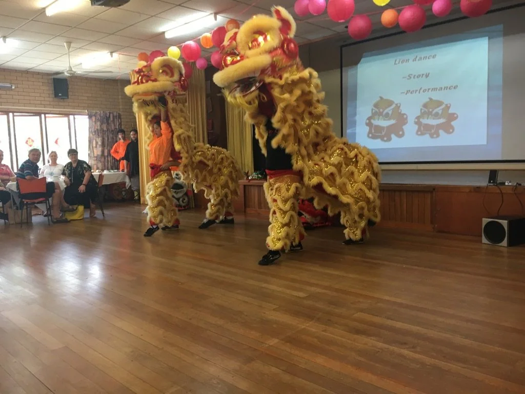 Two traditional Chinese lion dance performers in yellow costumes with red accents performing on a wooden floor, with an audience seated at tables and a projector screen displaying text in the background.