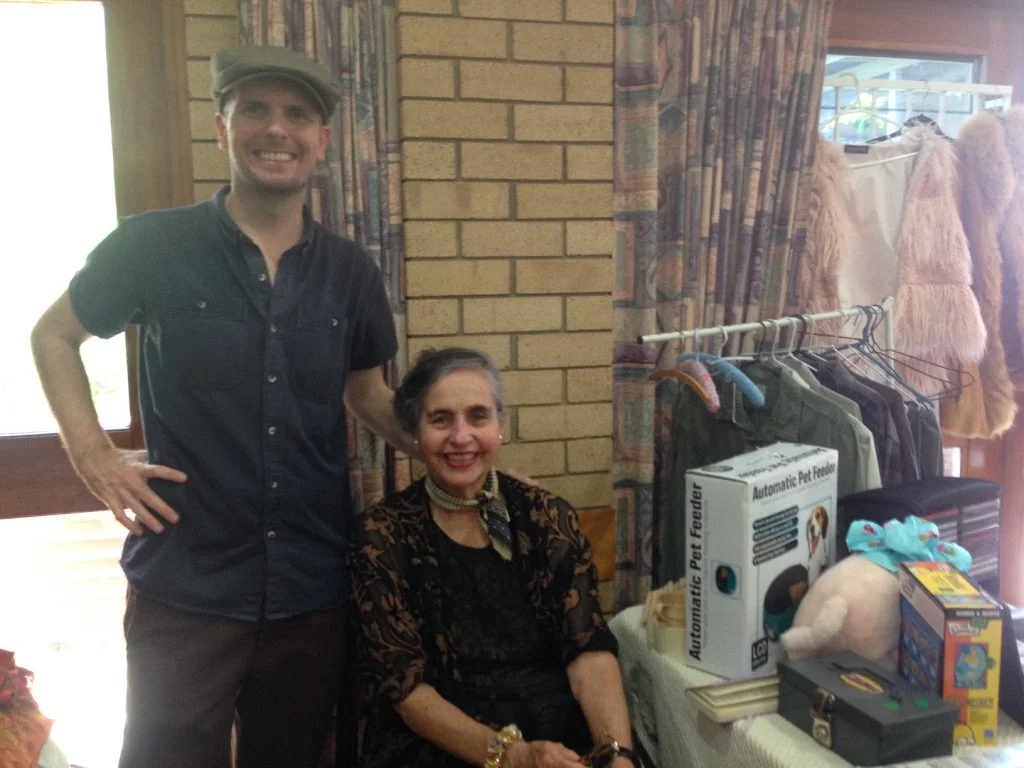 Two people, a man and a woman, are smiling and posing indoors, with clothing and pet supplies behind them.