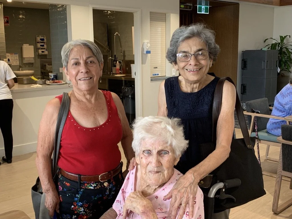 Three women, including an elderly woman in a pink shirt, are smiling and posing together in what appears to be a community or nursing home dining area. Two women stand behind the elderly woman, one wearing a red top and the other in a dark sleeveless