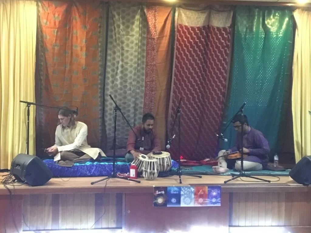 Three musicians sitting on a stage with colorful fabric backdrop, playing traditional instruments, microphones set up in front of them.