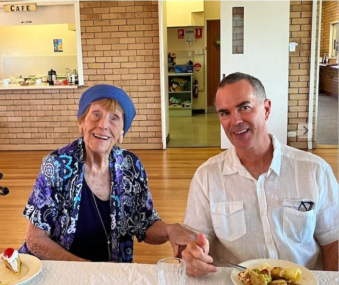 An elderly woman with a blue headscarf and a man sitting at a table, smiling and shaking hands, with a cake and a plate of food in front of them in a dining area.