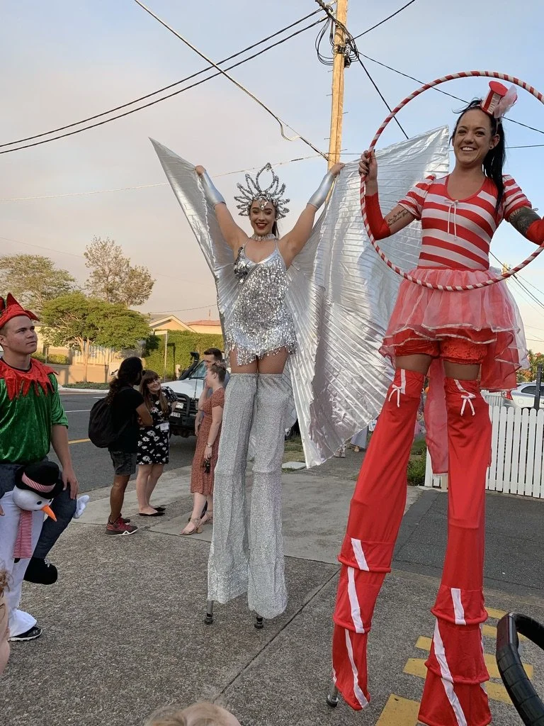 Two women on stilts dressed in festive costumes at an outdoor event, one with a silver sparkly costume with wings and a crown, and the other in a red and white striped outfit with a hoop, surrounded by onlookers.