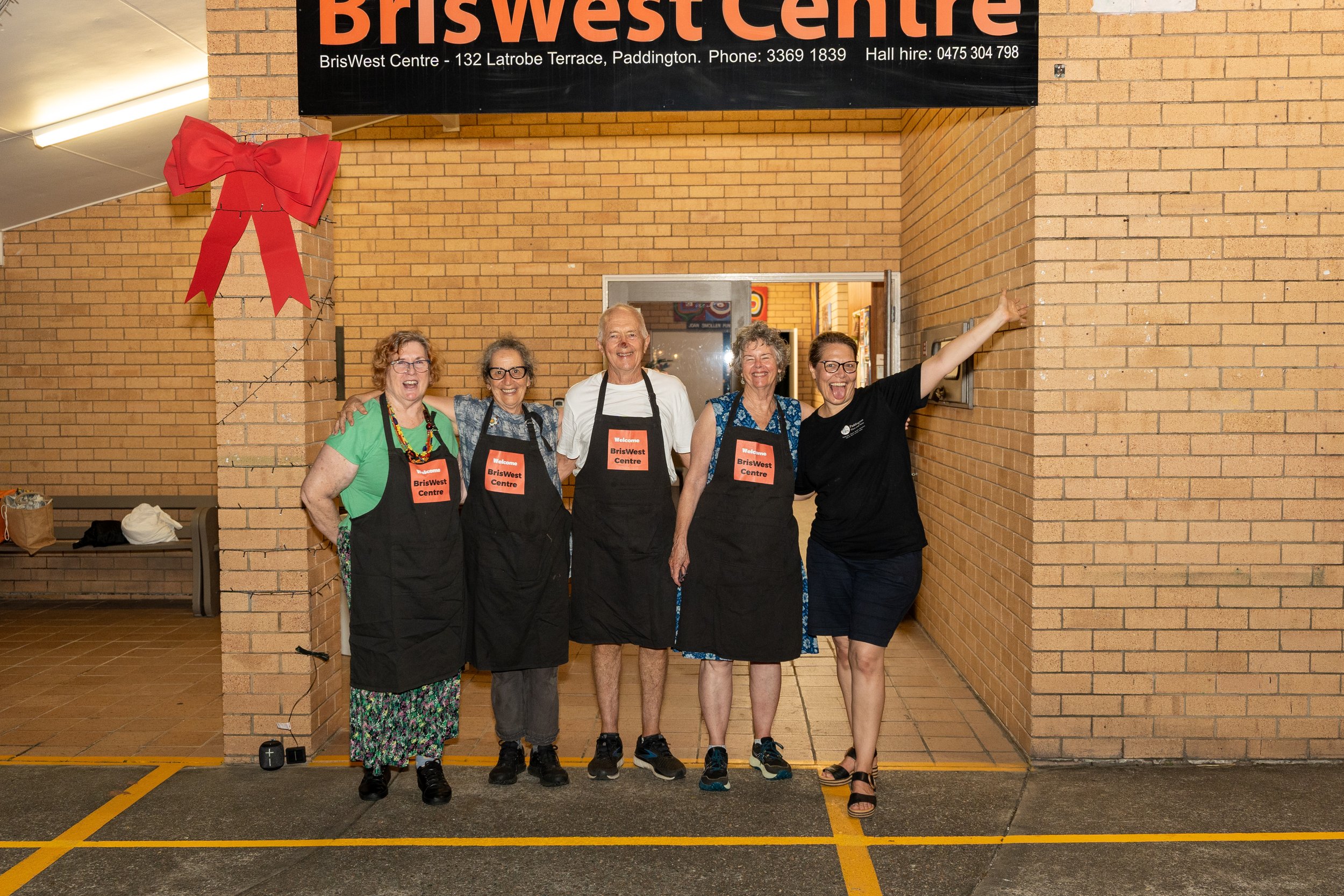 Five women standing together in front of a brick wall, smiling and wearing aprons with a BrisWest Centre badge, celebrating a community event.