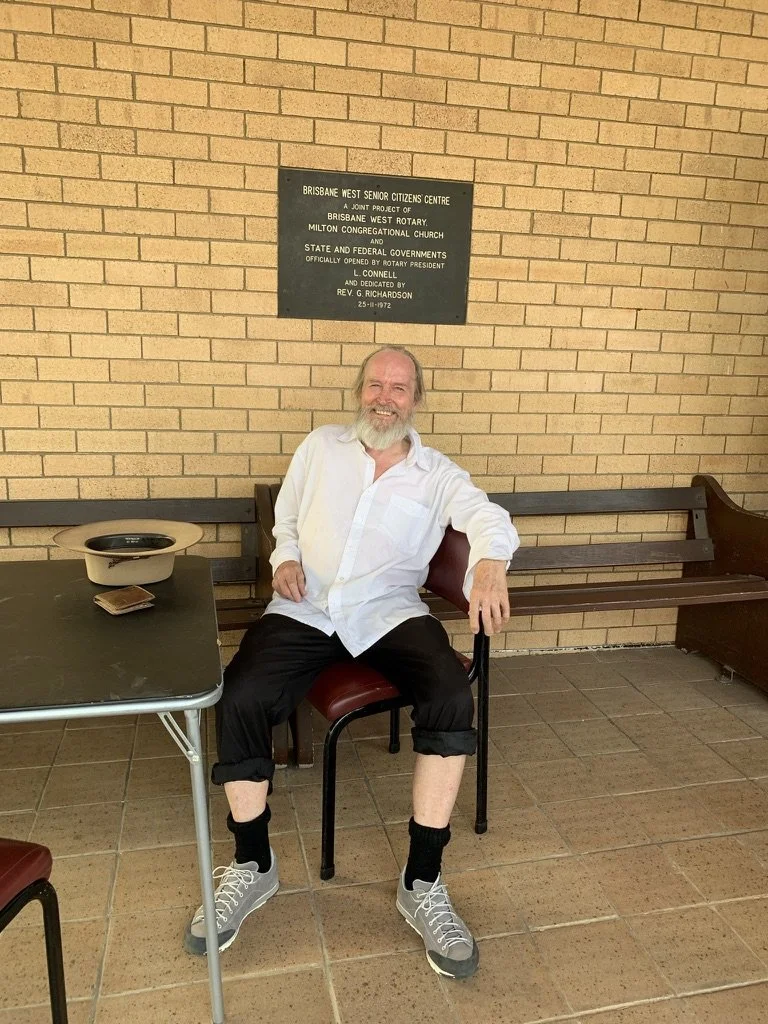A man with a beard wearing a white shirt, black pants rolled up to the ankles, black socks, and gray sneakers sitting on a chair in front of a brick wall inside a building.