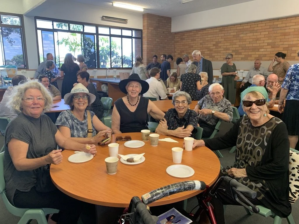 Six elderly women sitting around a table with cups and plates, smiling at a social gathering in a well-lit room with large windows and a brick wall, other people standing and sitting in the background.