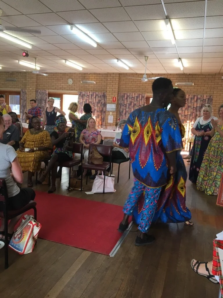 People sitting and standing in a community room, some watching and others taking photos, with two dancers dressed in colorful traditional African attire performing in the foreground.