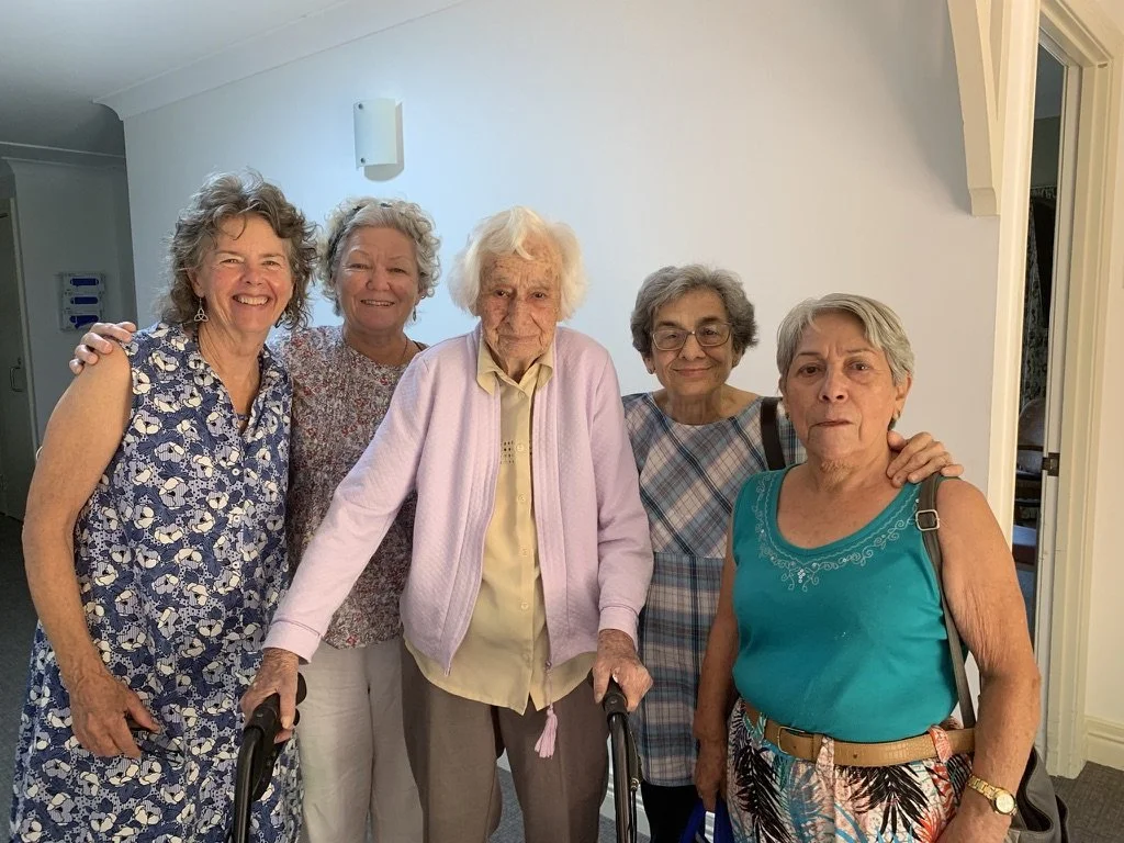 Five elderly women standing together indoors, smiling, with one using a walker.