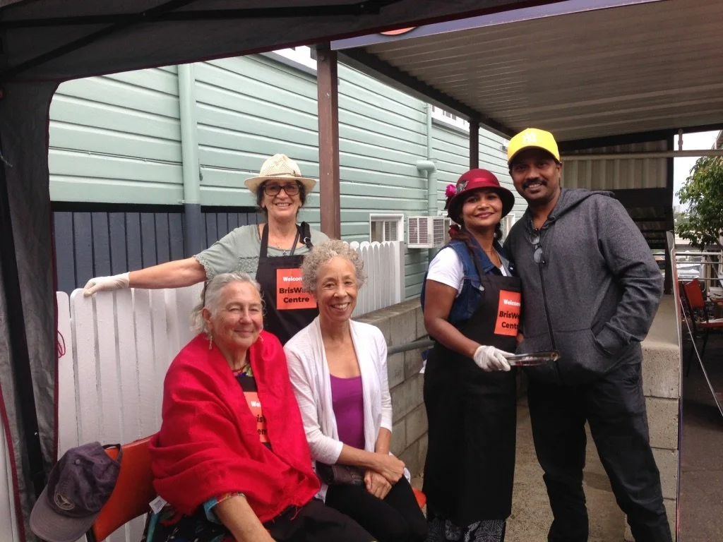 Five people, four women and one man, are gathered under a canopy at an outdoor event. Two women are seated, one wrapped in a red shawl and smiling, the other in a white cardigan. Two women are standing, wearing aprons and hats, and a man in a gray ho
