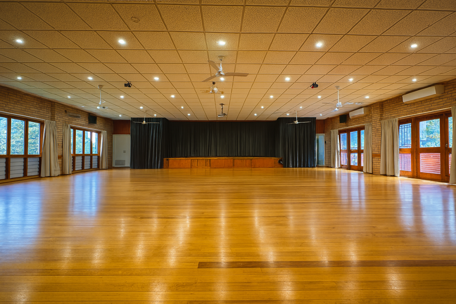 An empty wooden-floor banquet hall with windows on both sides and a stage at the front, decorated with dark curtains.