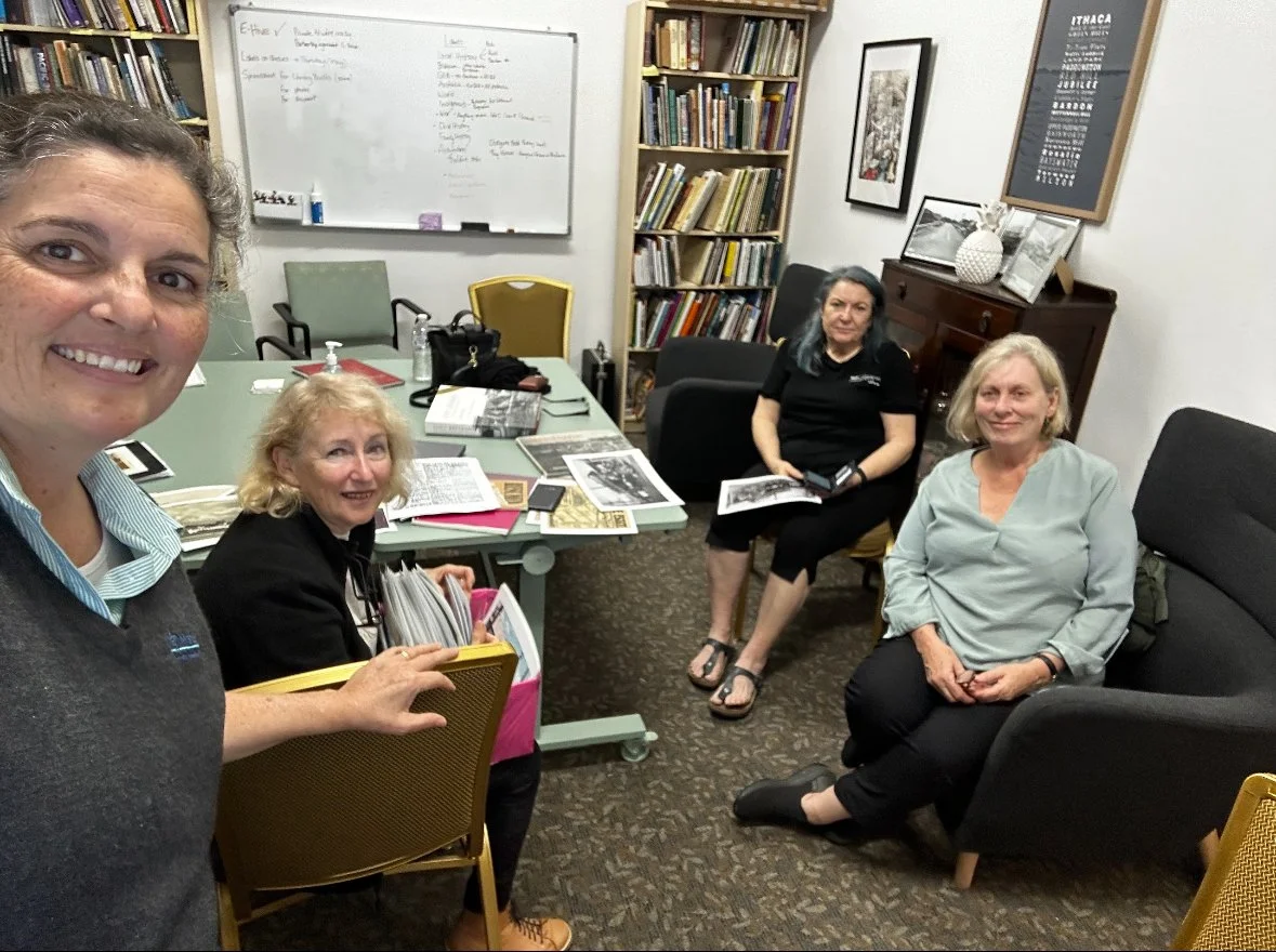 Four women sitting and standing in a library or office room, with bookshelves, framed pictures, and a whiteboard in the background, some reading or looking at papers