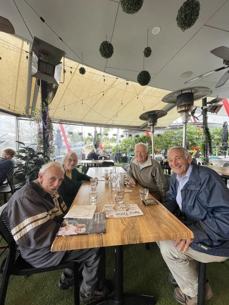 Four elderly people sitting at a wooden table inside a restaurant or cafe, smiling and enjoying drinks. The setting is decorated with hanging green plants and outdoor heaters, with natural light coming through large windows in the background.