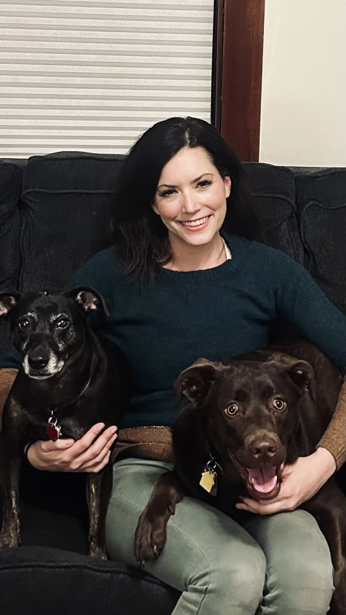 A woman with black hair smiling and sitting on a black couch with two dogs, one black and one brown, both looking at the camera.